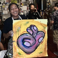 a woman smiles while holding up a painting in a studio